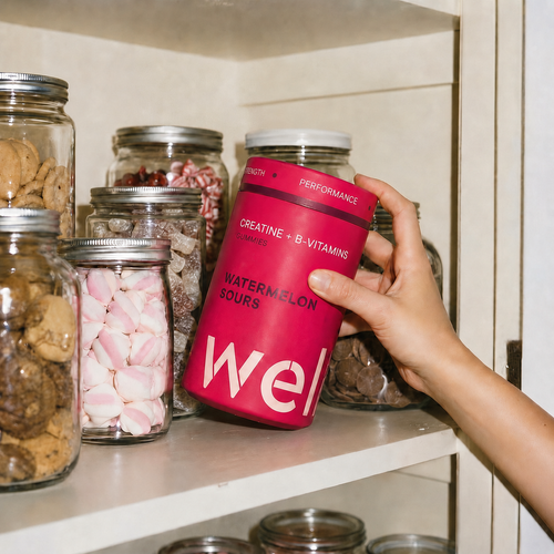 Hand reaching for a tub of WELLF Watermelon Sours creatine gummies on a pantry shelf lined with jars of confectionery and sweet treats